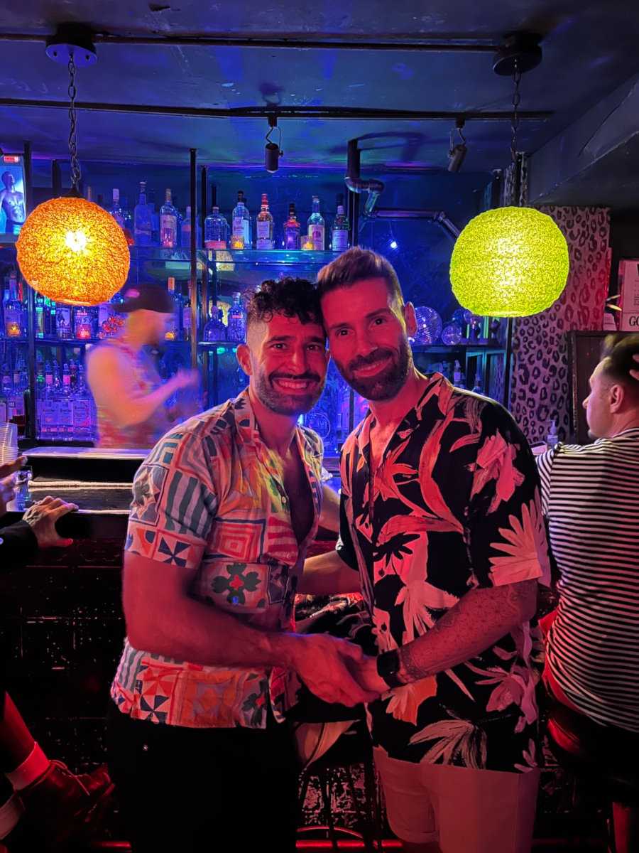 Seby and Stefan holding hands and smiling at the camera while standing in front of the bar at Barracuda lounge in New York City.