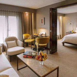 A plush hotel sitting room with chairs and a coffee table leading into a bedroom.